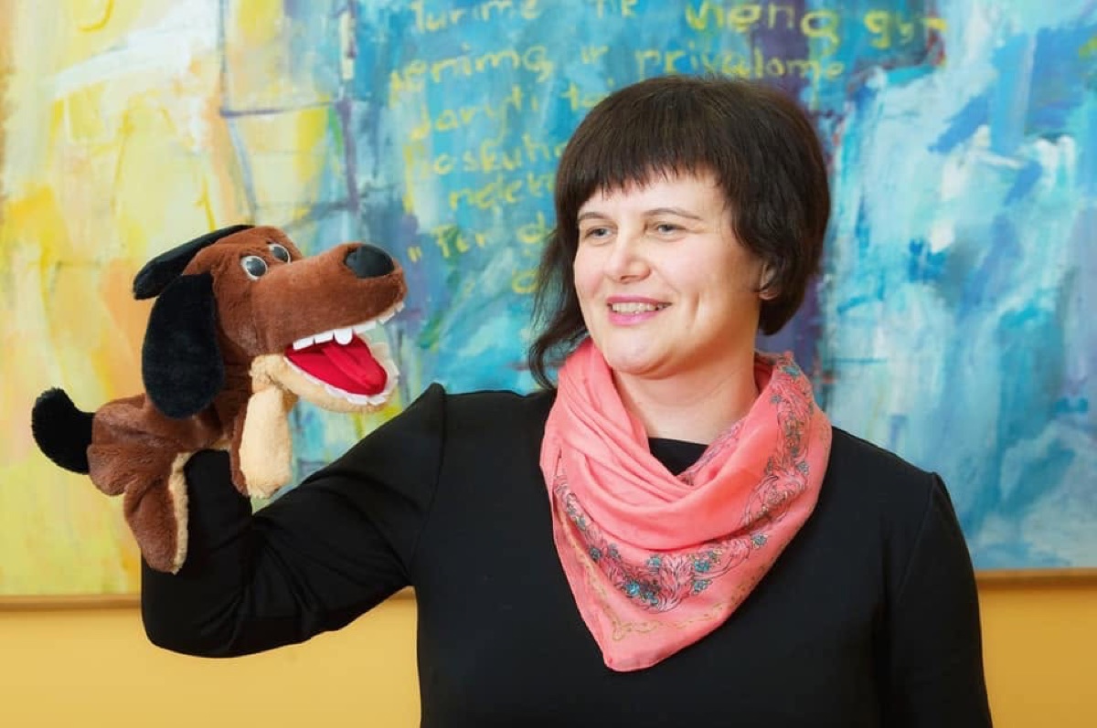 Logopedė Diana su šuns pirštinine lėle prie spalvingo paveikslo